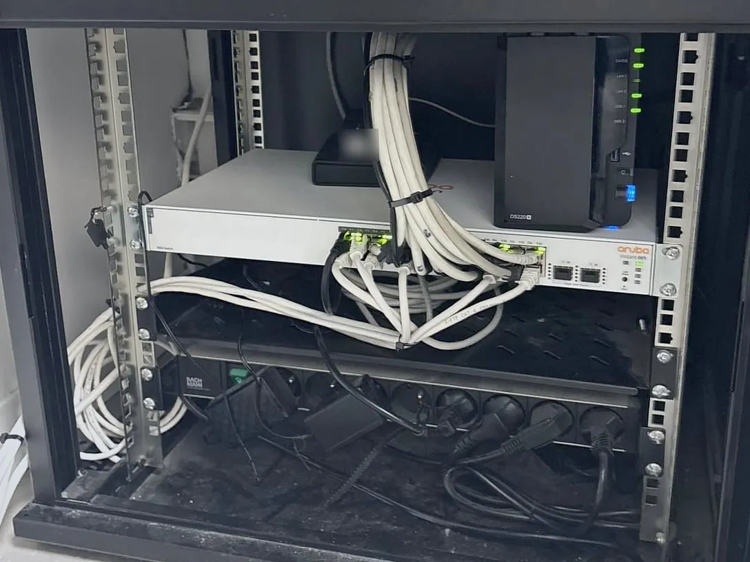 Baie réseau rangée : câbles blancs réunis en faisceau par des colliers jusqu’au switch Aruba, NAS Synology à côté, bloc de prises en bas