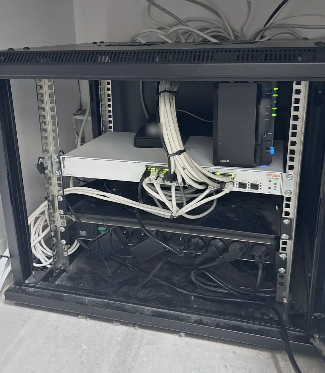 La même baie rangée : câbles blancs réunis en faisceau par des colliers jusqu’au switch Aruba, NAS Synology à côté, bloc de prises en bas