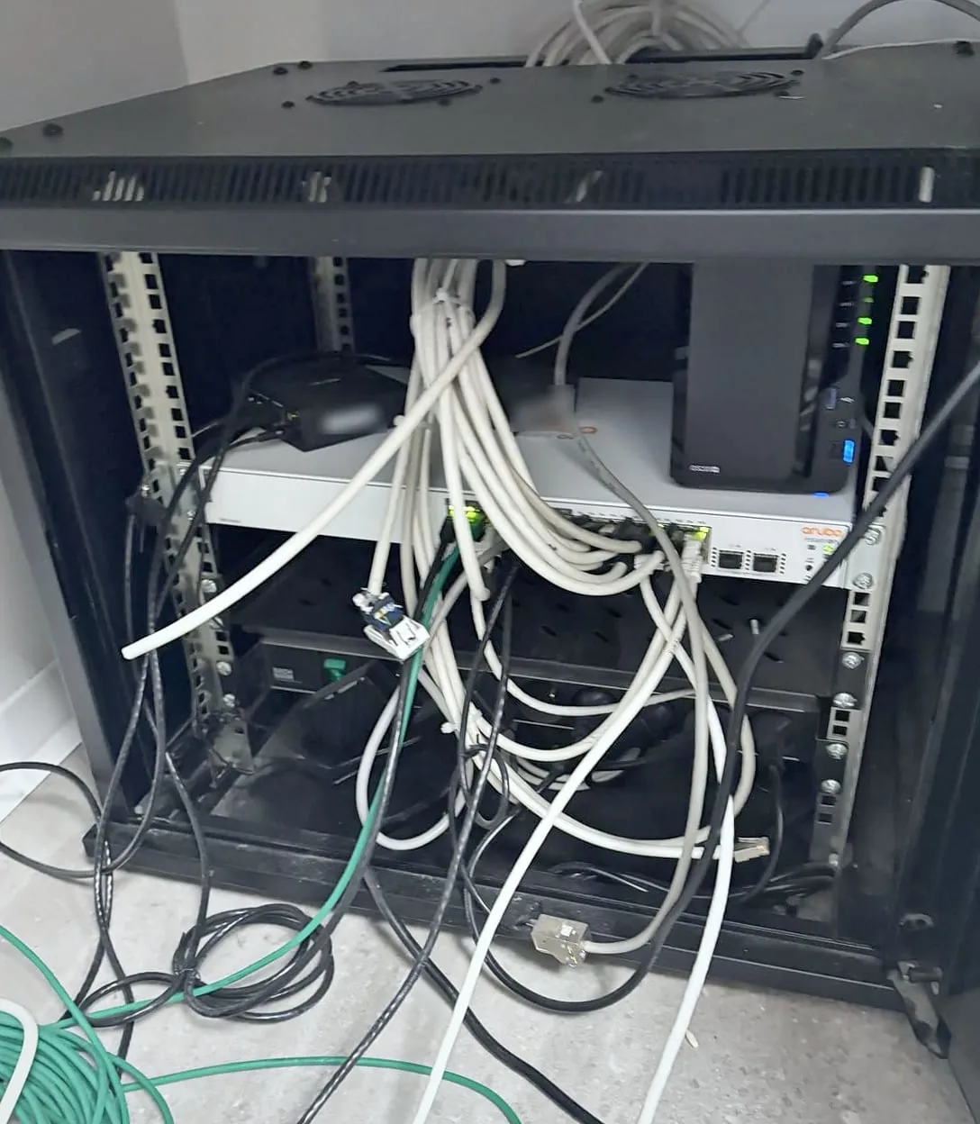 Baie réseau noire ouverte, câbles blancs emmêlés qui pendent devant un switch Aruba et un NAS Synology, bobine de câble vert au sol