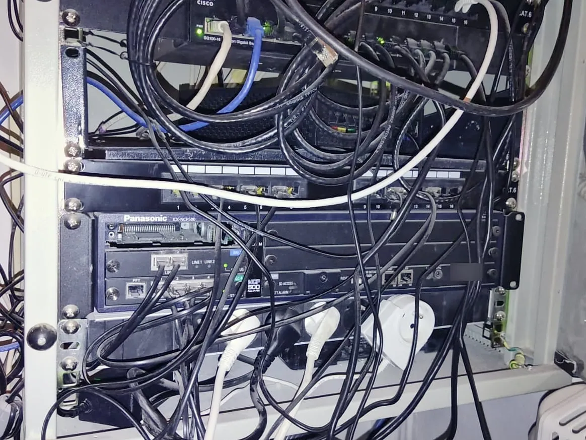Baie réseau encombrée de câbles noirs, blancs et bleus emmêlés devant un switch Cisco et un standard téléphonique Panasonic