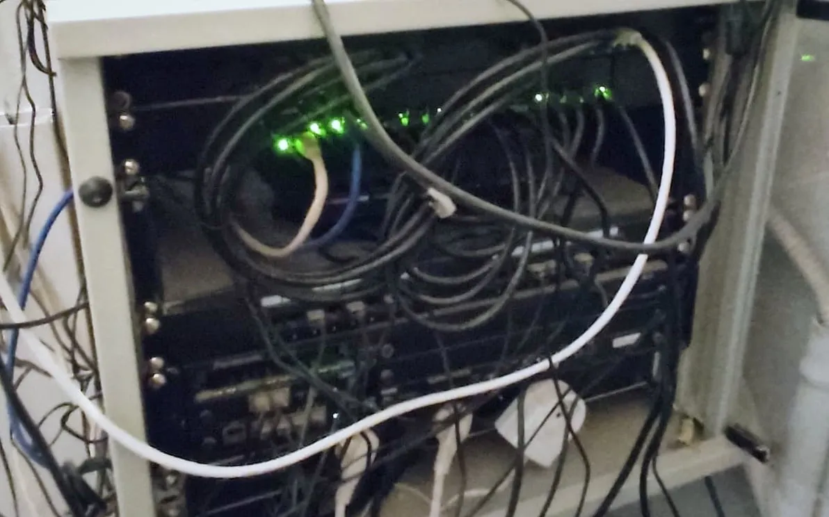 Petite baie réseau sous une étagère, câbles noirs et blancs emmêlés devant un switch aux voyants verts