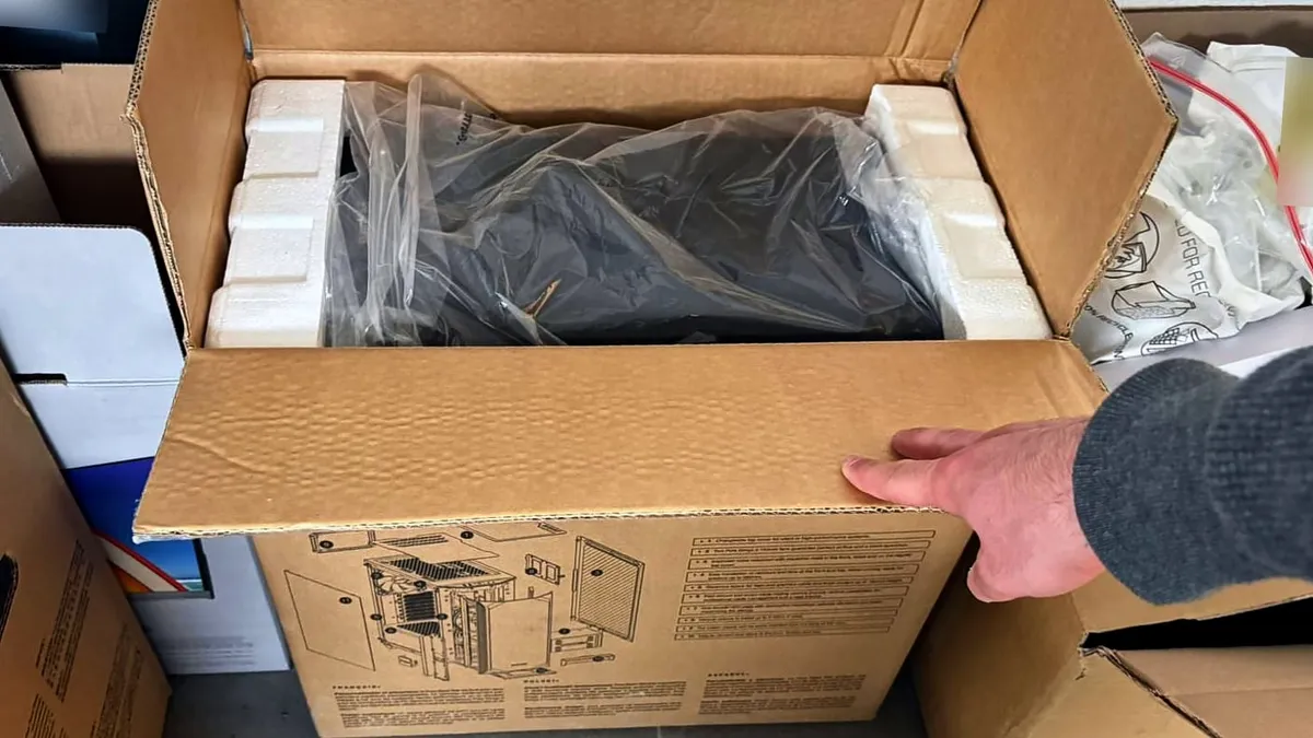 Carton ouvert, boîtier noir sous plastique entre ses cales, une main tient le rabat