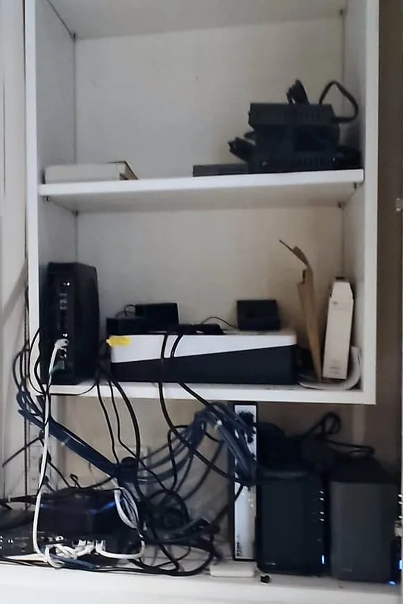 Placard technique ouvert avec des câbles réseau emmêlés, une box et un switch posés en vrac sur l’étagère du bas